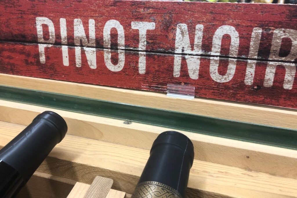 The necks of two wine bottles placed in a rack, a rustic wooden sign with a red painted background and the words Pinot Noir printed on in white.